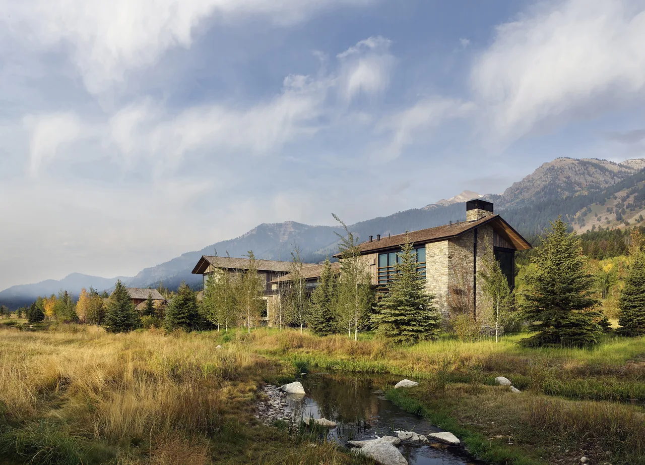 Stone and wood house with large windows beside a small creek with trees and mountains in the background.