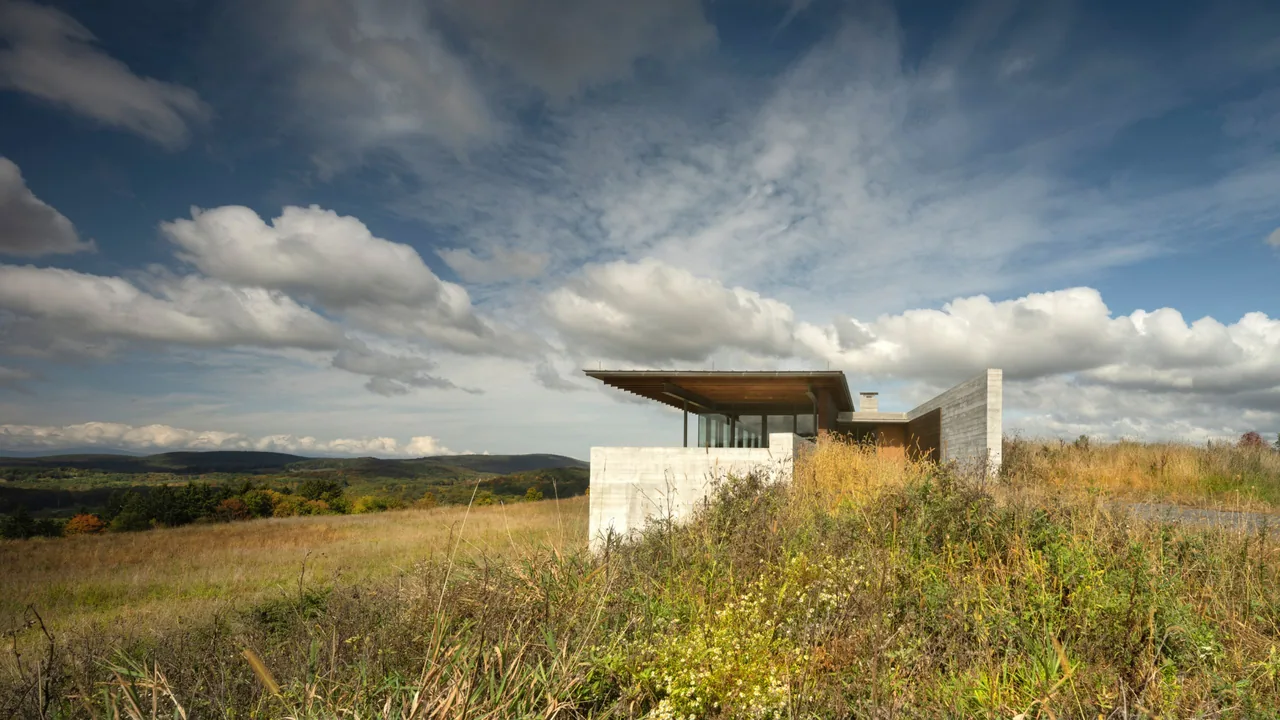 Modern house with flat roof and concrete walls in a grassy landscape under a partly cloudy sky.