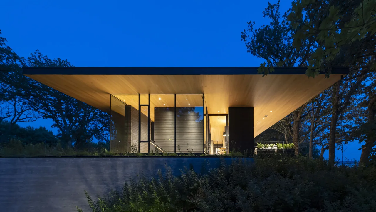 Night view of a modern house with an extensive overhanging flat roof and glass walls surrounded by trees.