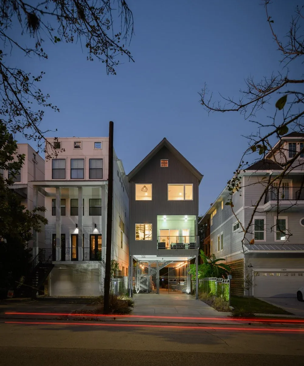 Evening view of Saunders House featuring illuminated modern three-story home elevated on stilts with balcony and large windows.