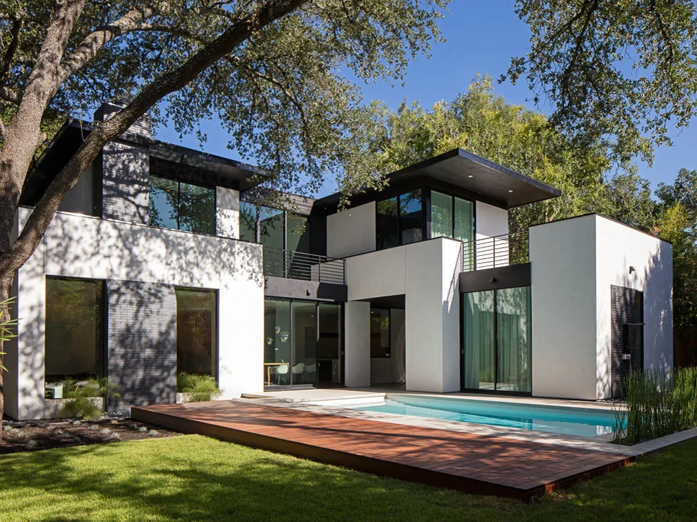 Modern two-story home with white stucco exterior, flat roofs, large windows, and a pool with an adjacent wood deck.