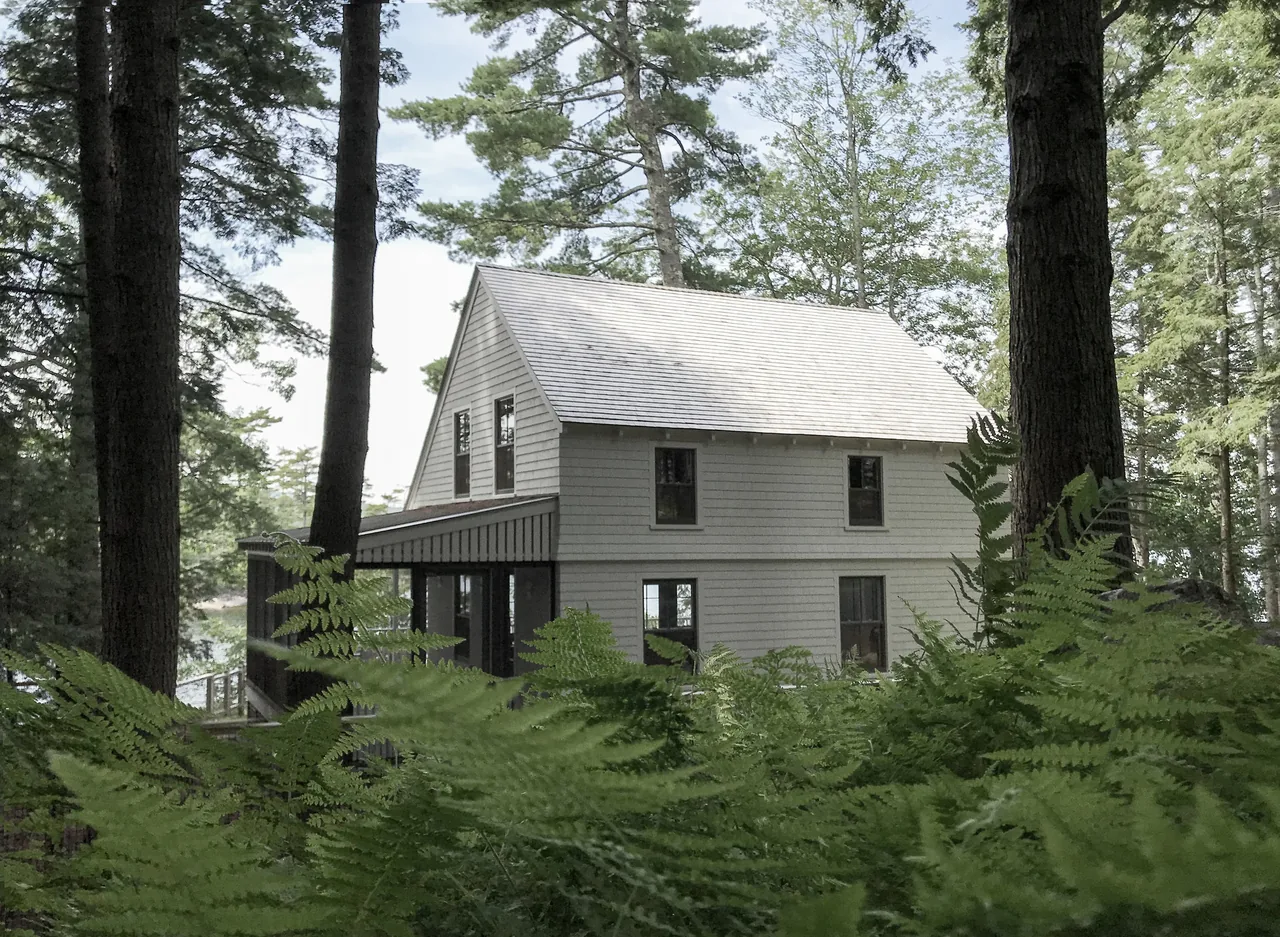 A two-story cottage with white shingle siding and a steep gable roof, surrounded by tall trees and lush ferns.
