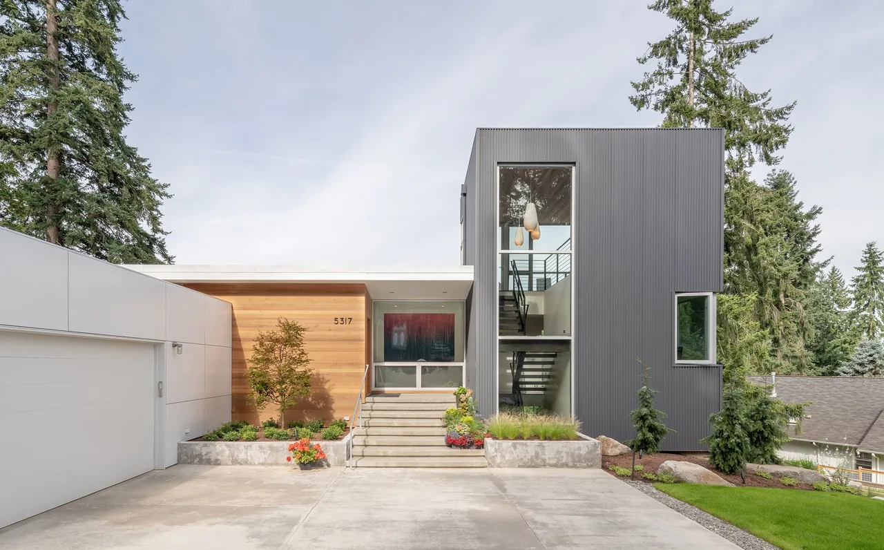 Front view of a modern two-story house with gray metal siding, wood paneling, and large vertical windows, surrounded by trees.