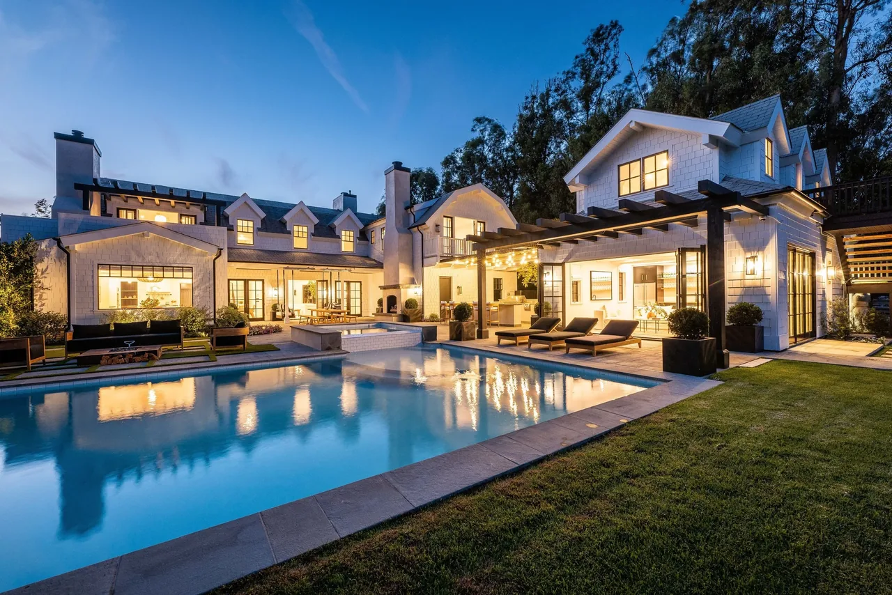 Evening view of a traditional-style two-story house with illuminated windows, a swimming pool, and outdoor lounge area.