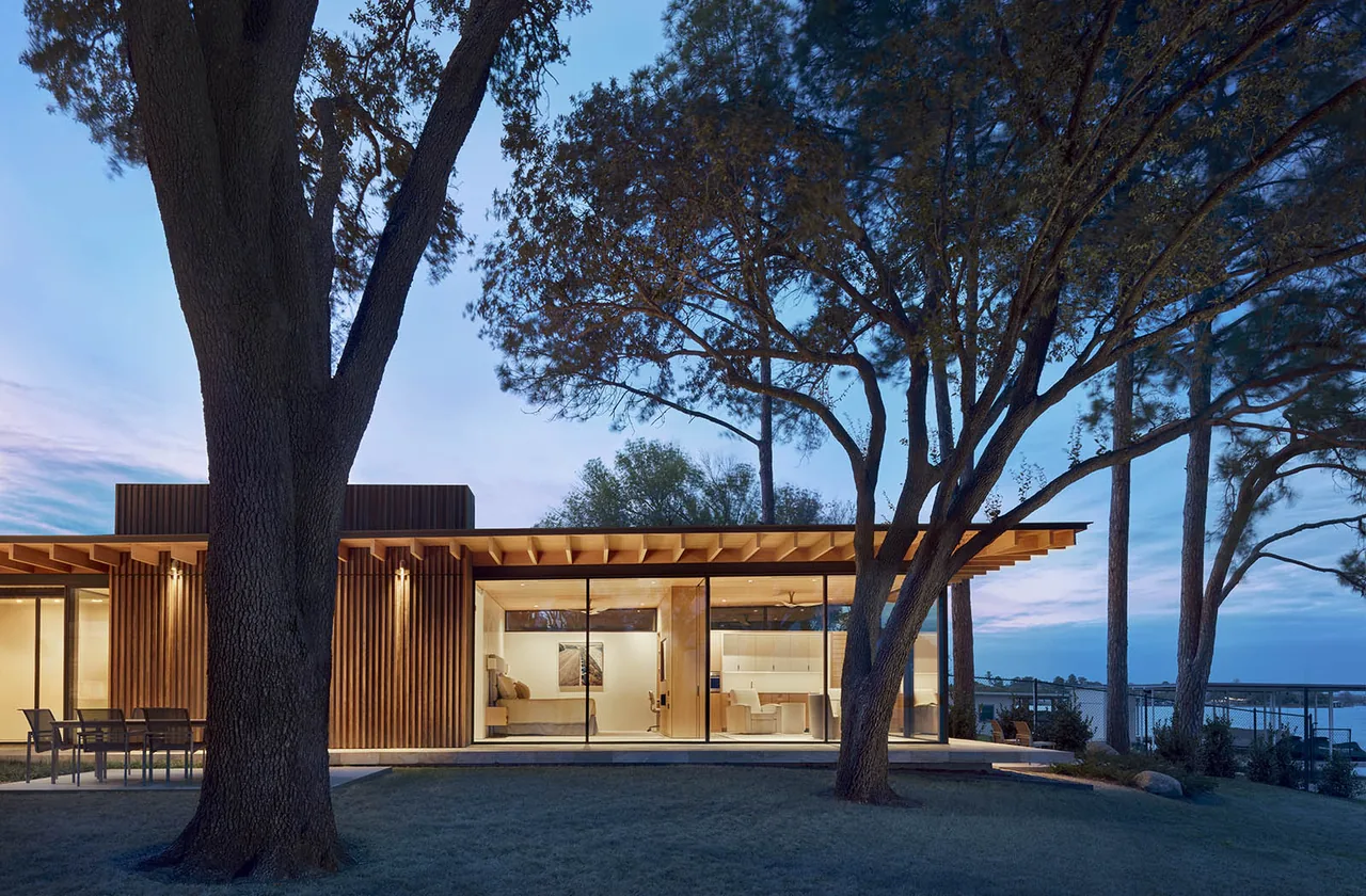 View of a modern single-story lakeside retreat with large glass sliding doors and a flat roof, framed by large mature trees at dusk.