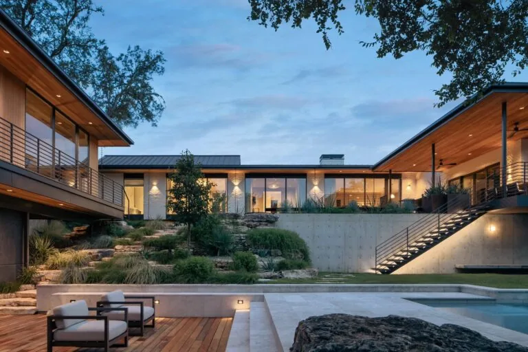 Evening view of a modern U-shaped home with a pool, elevated concrete patio, and landscaped yard in front.