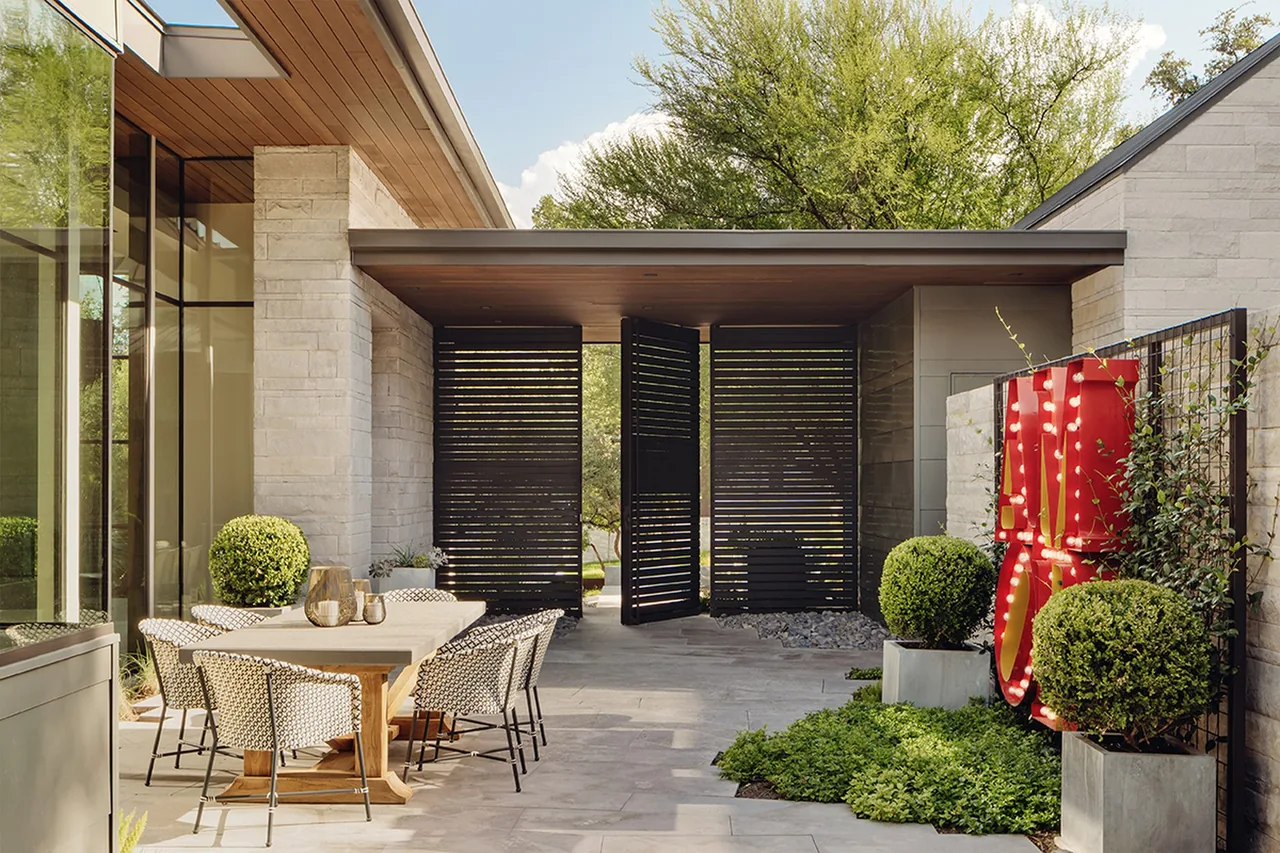 Outdoor dining area with a wooden table and patterned chairs next to modern stone and glass architecture under a roofed walkway with slatted