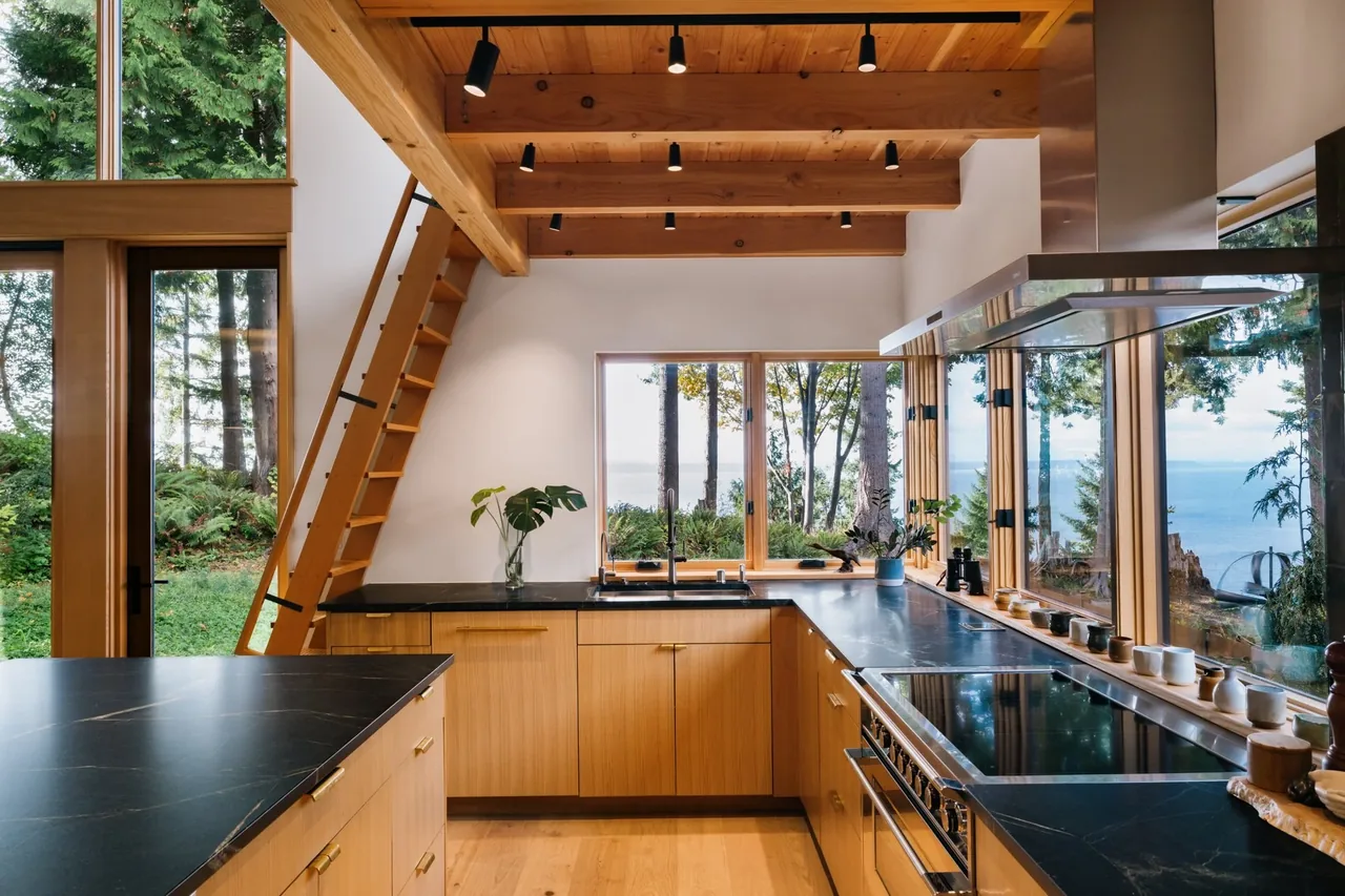 Interior view of a kitchen with wood cabinetry, black countertops, and large windows showing a forest and ocean view.