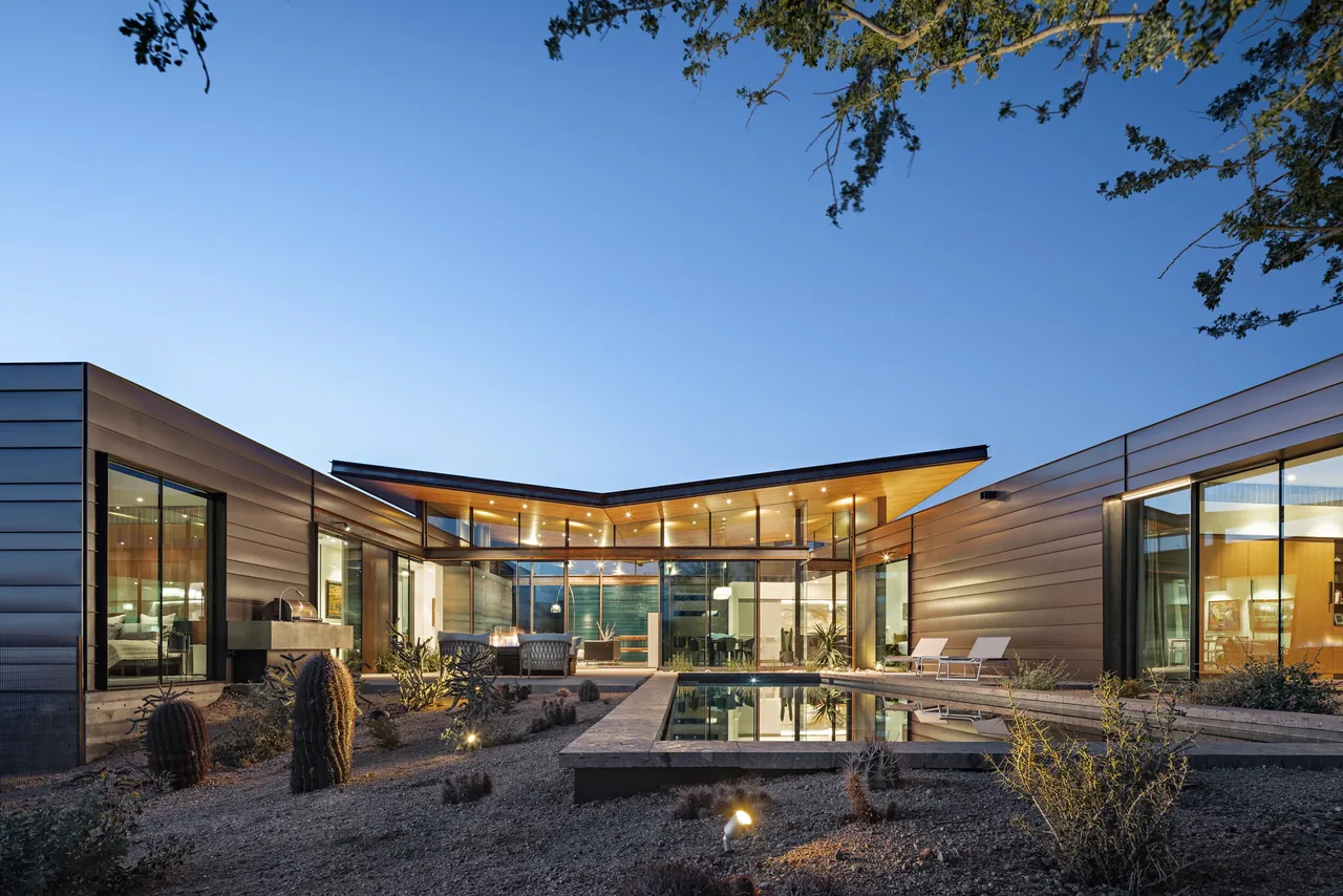 A modern one-story residential building with a flat roof, large glass windows, and an outdoor pool surrounded by desert landscaping.
