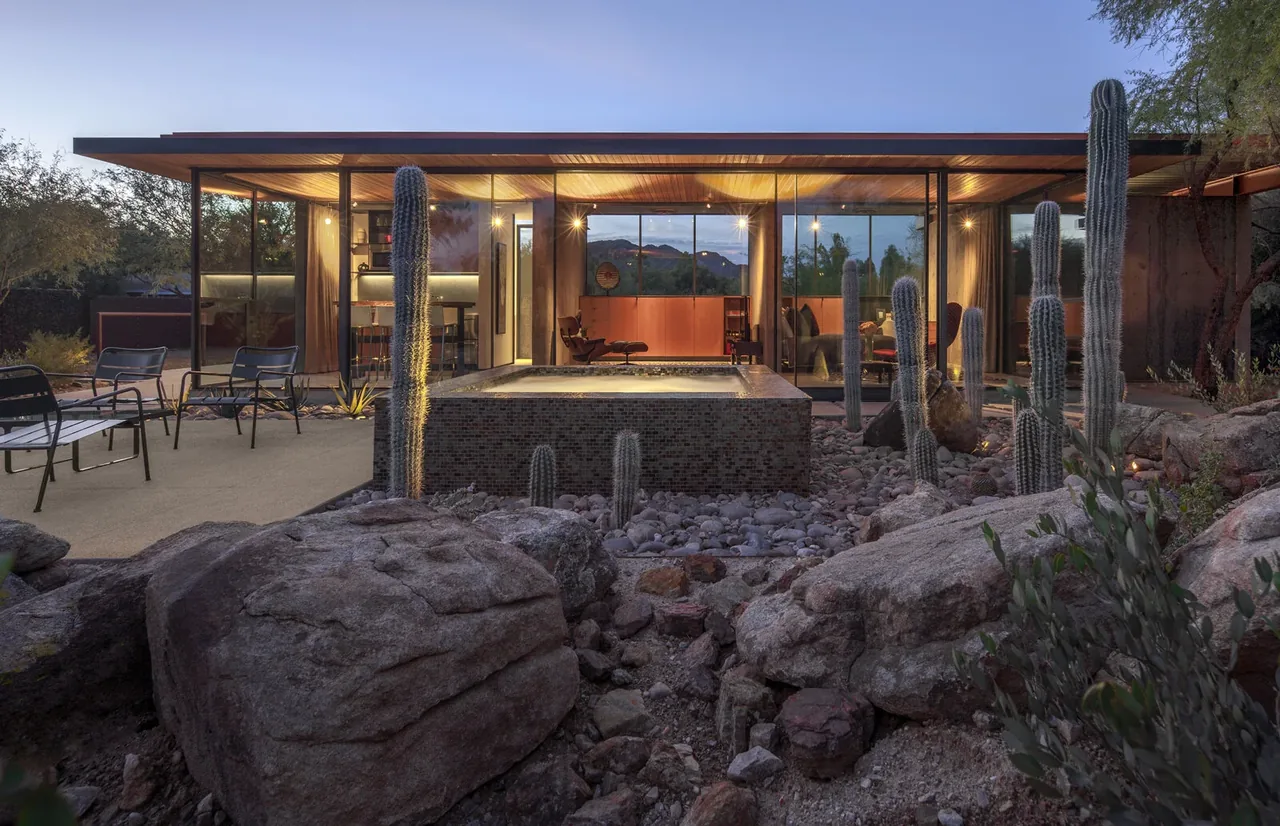 Evening view of a modern barn guest house with large glass walls and a reflecting pool surrounded by desert landscape.