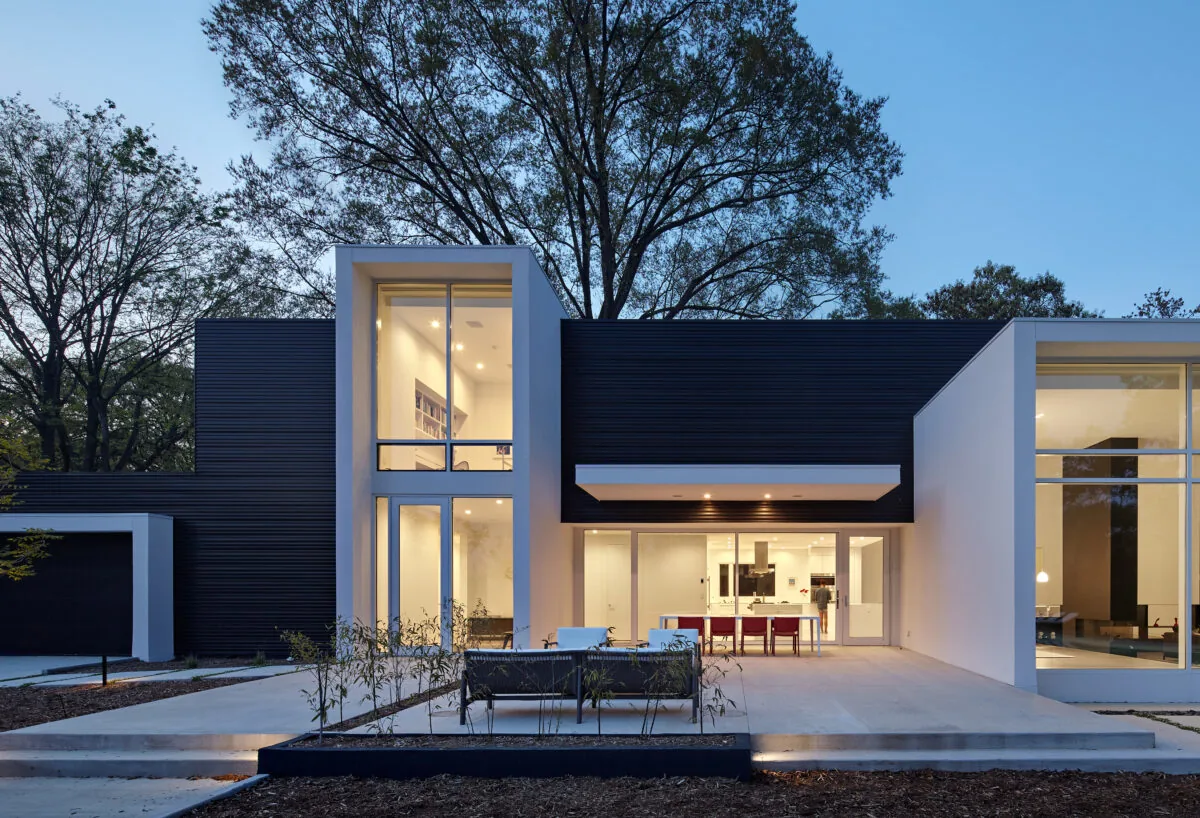 Evening view of a modern two-story house with large vertical windows and a dark exterior frame contrasted with white walls.