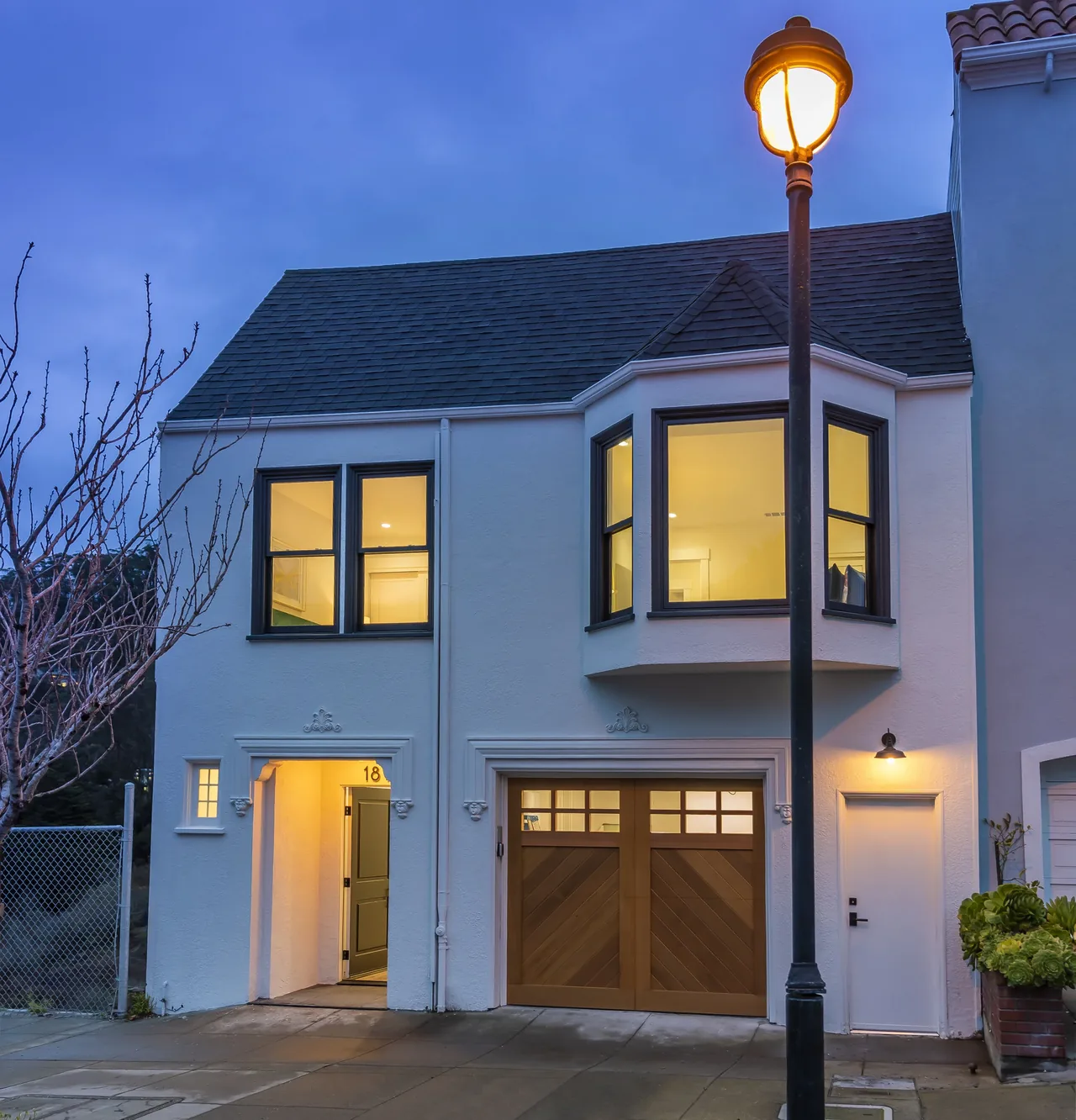 Two-story white stucco house with dark roof and wood-paneled garage door at dusk with a street lamp in front.