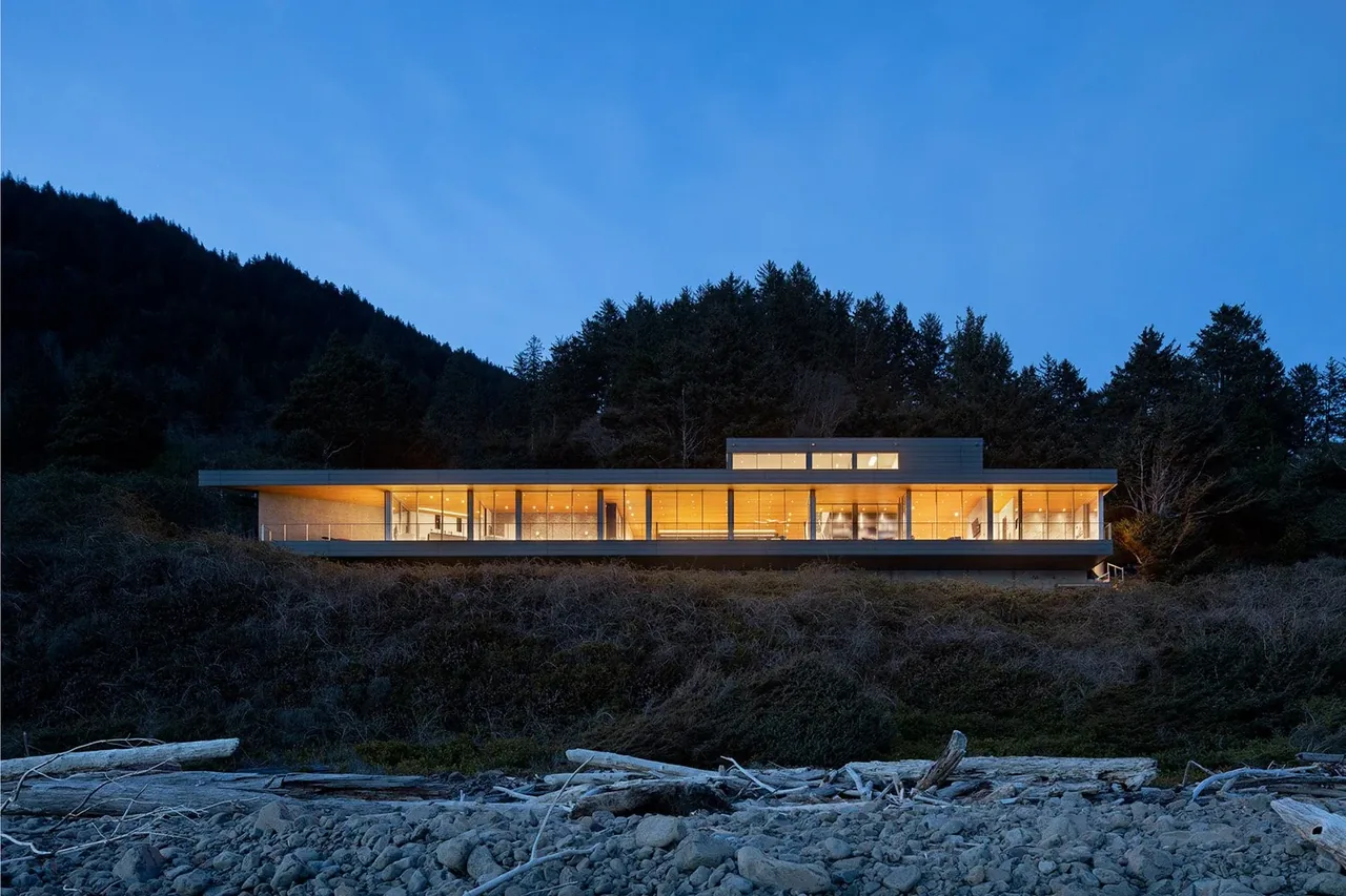 Long, low modern house with floor-to-ceiling glass illuminated at dusk near a forested hillside.
