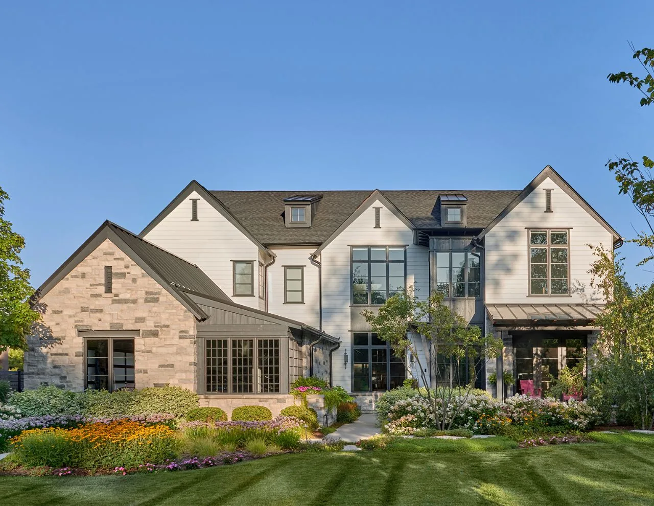 Front exterior view of a two-story transitional farmhouse residence with stone and white siding under a clear blue sky.
