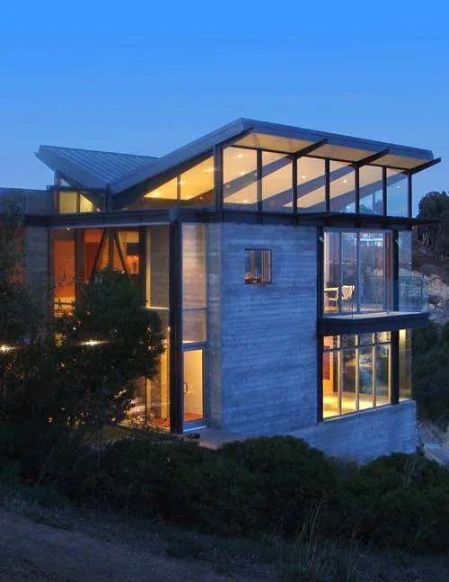 Night view of a modern house with large glass windows and an angled flat metal roof on a hillside.