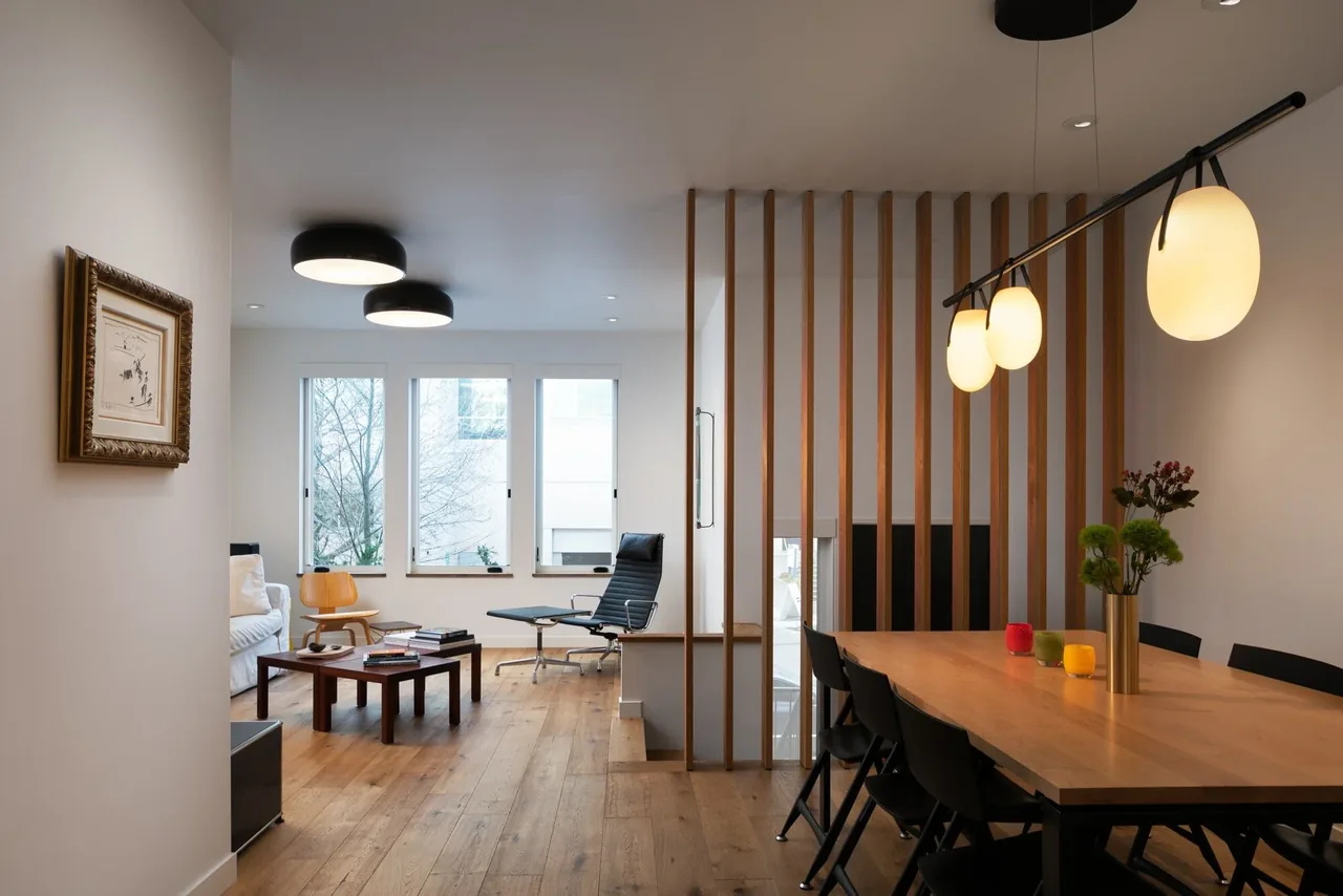 Modern townhouse interior with a dining area featuring hanging lights and a living space with wood flooring and large windows.