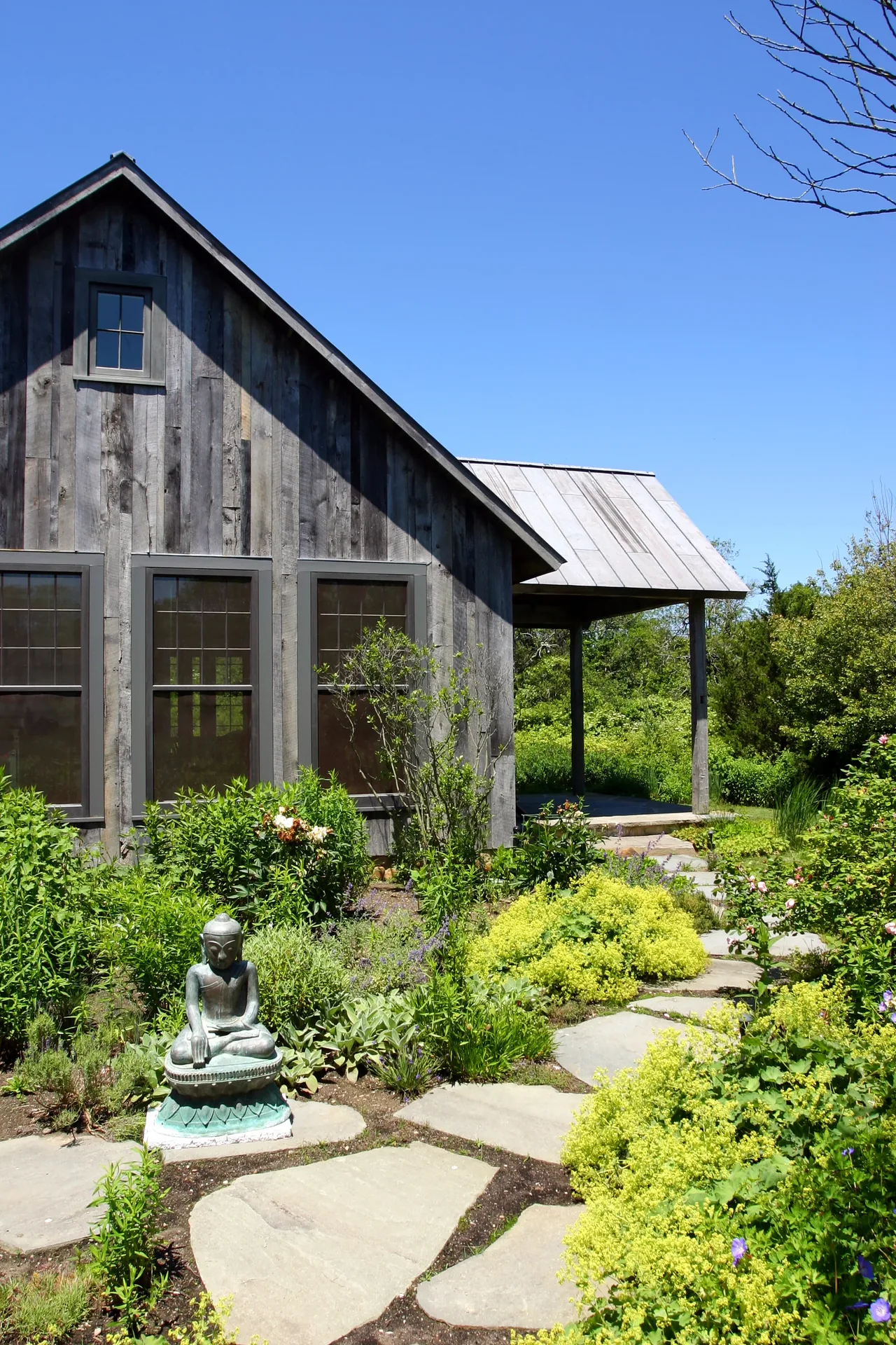 Exterior view of a rustic wooden guest cottage with a stone pathway and lush garden featuring a Buddha statue.