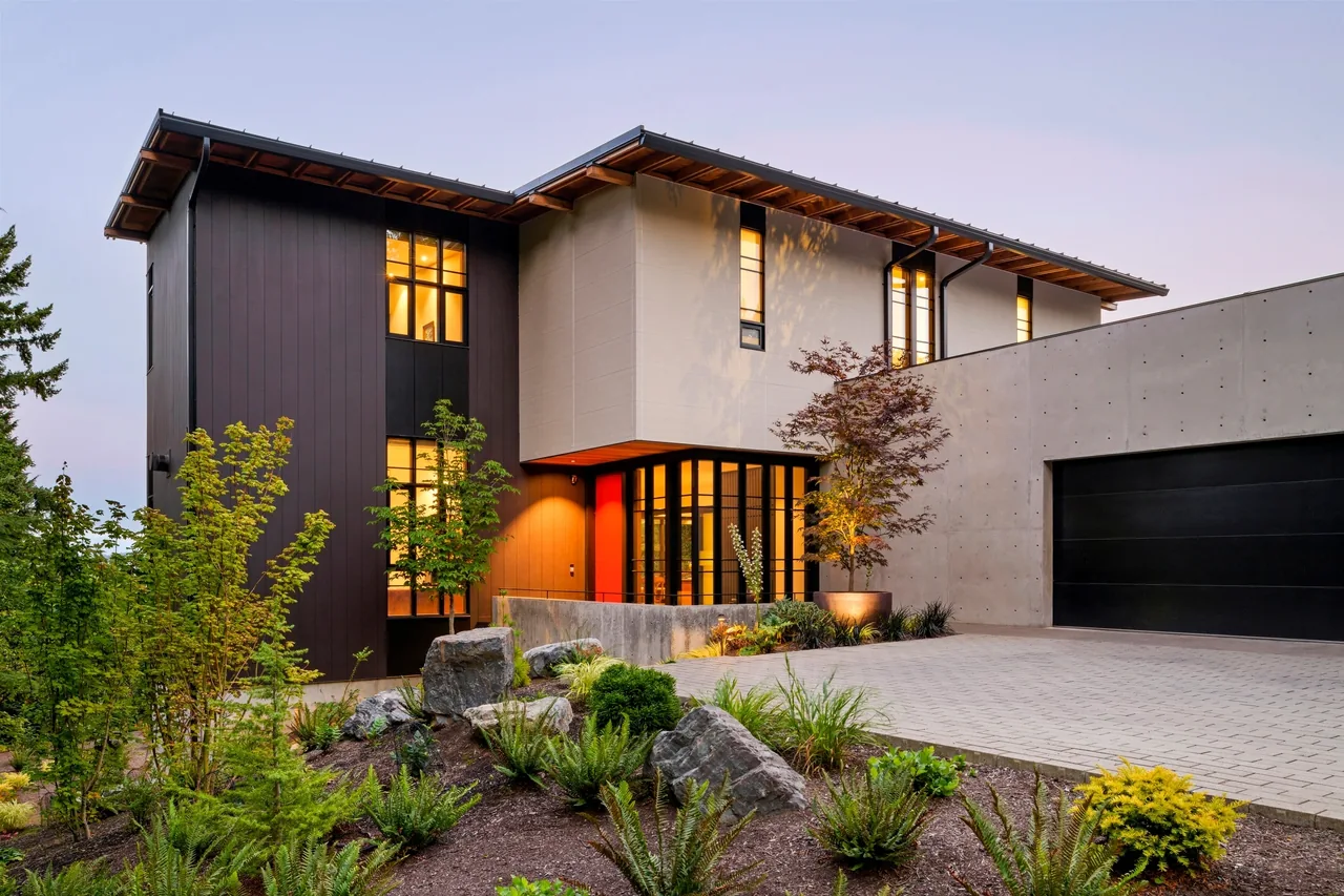 A modern two-story house with dark vertical siding, light stucco, concrete garage wall, and landscaped front yard with rocks and plants at d