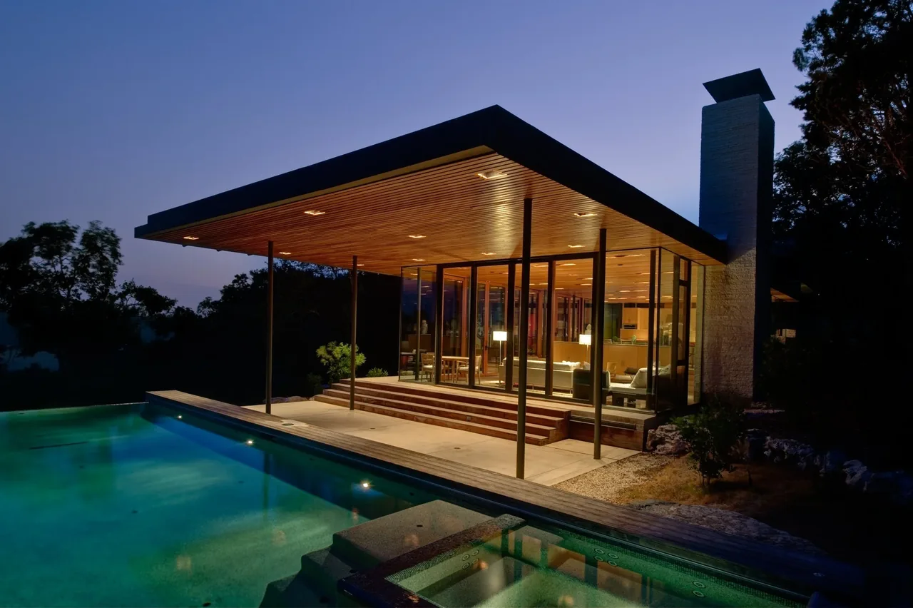 Night view of a modern house with a wooden ceiling and large glass walls next to a lit swimming pool and spa.