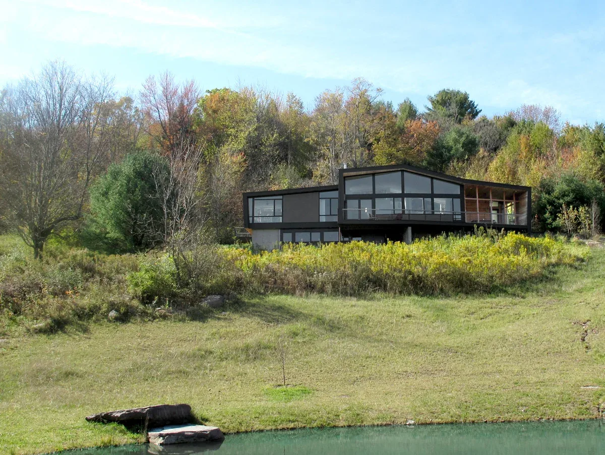 Black modern house with large windows sits on a hillside surrounded by trees and overlooks a small pond.