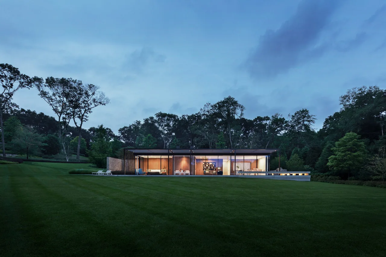 A modern pavilion with large glass walls set on a wide green lawn surrounded by trees at dusk.