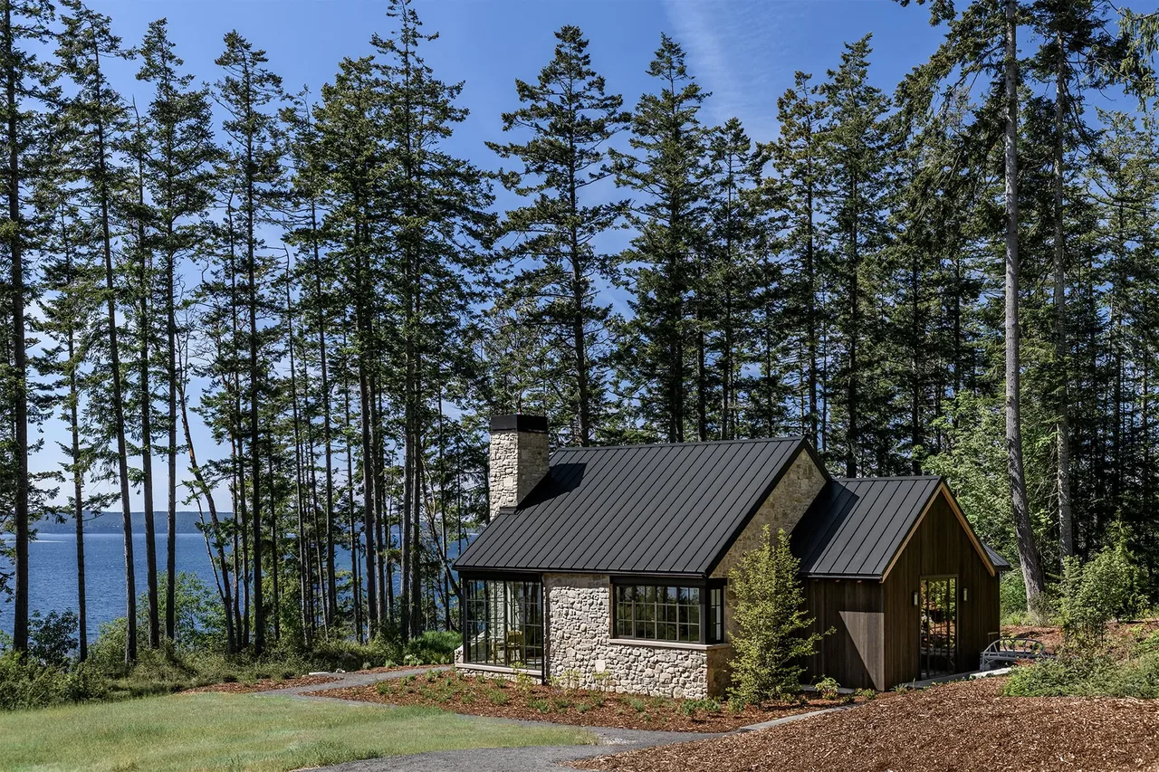 A cottage with stone and wood siding and a black standing seam metal roof in a forested area near a lake.