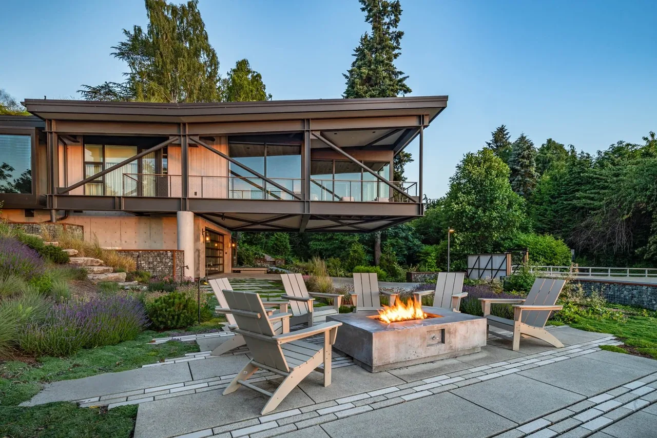 Modern cantilevered house overlooking an outdoor seating area with a fire pit surrounded by wooden chairs.