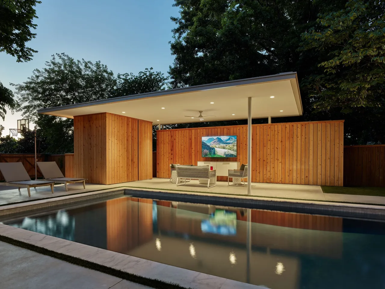 A modern poolside pavilion with wood siding, flat roof, and outdoor seating near a swimming pool at dusk.