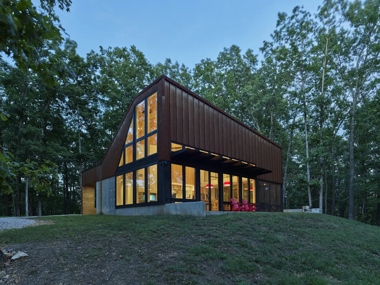 Exterior view of a modern two-story house with a rust-colored facade and large floor-to-ceiling windows surrounded by trees.