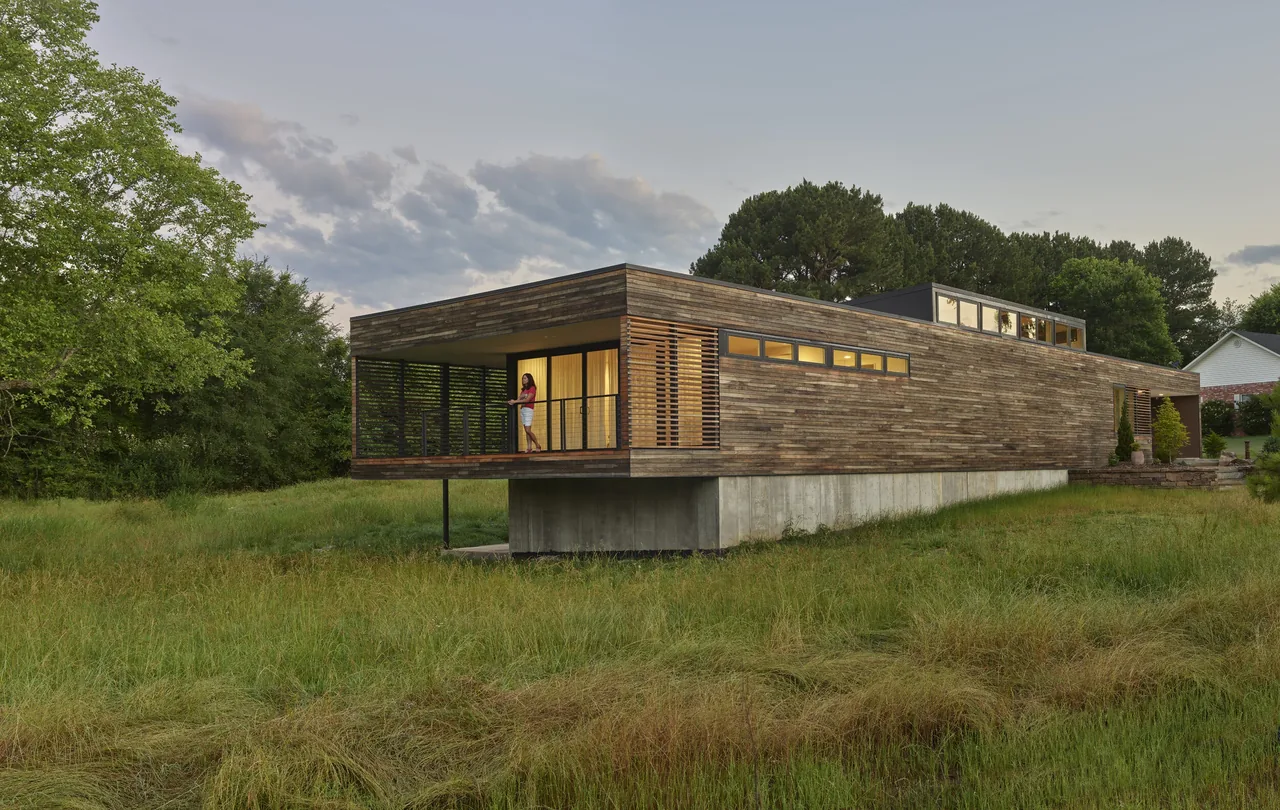 Modern house with horizontal wood siding elevated on a concrete base and featuring a covered balcony with wood slat details.