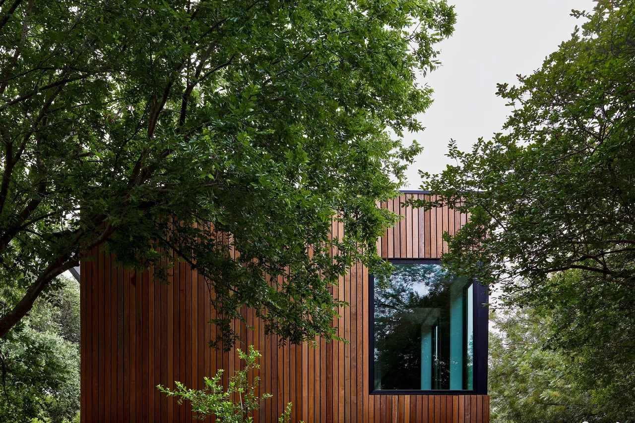 Exterior view of a modern wooden house with vertical wood siding and a large black-framed window partially obscured by green trees.