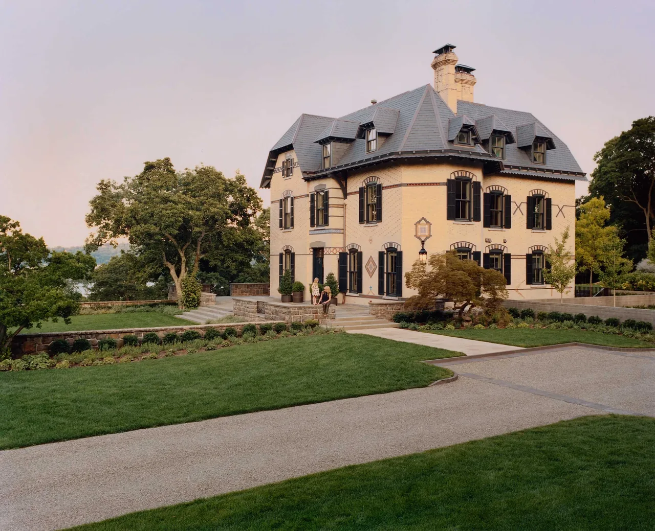 Brick estate house with multiple dormer windows and slate roofing surrounded by landscaped lawns and trees.