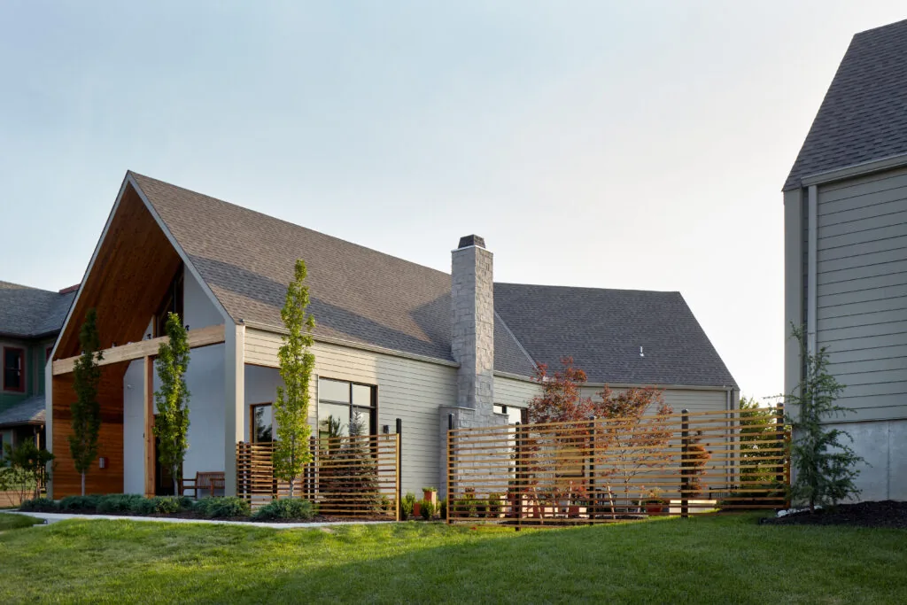 Exterior view of a modern house with a large gable roof, stone chimney, wooden fence, and green lawn under a clear sky.