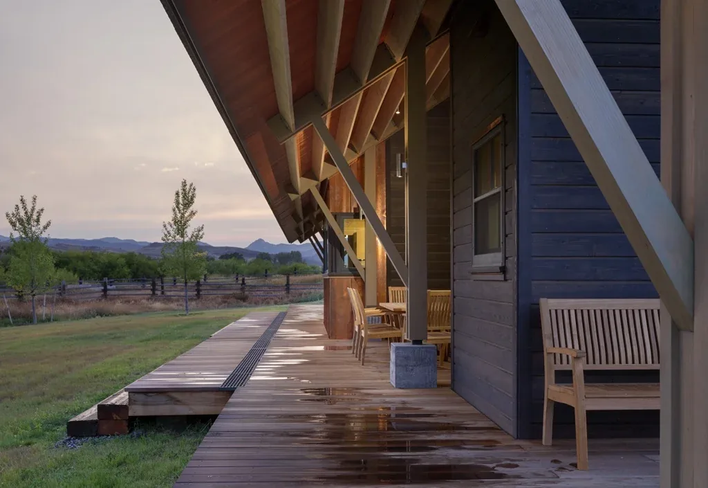 Wooden porch with slanted roof and outdoor seating overlooking grassy landscape and mountains at dusk.