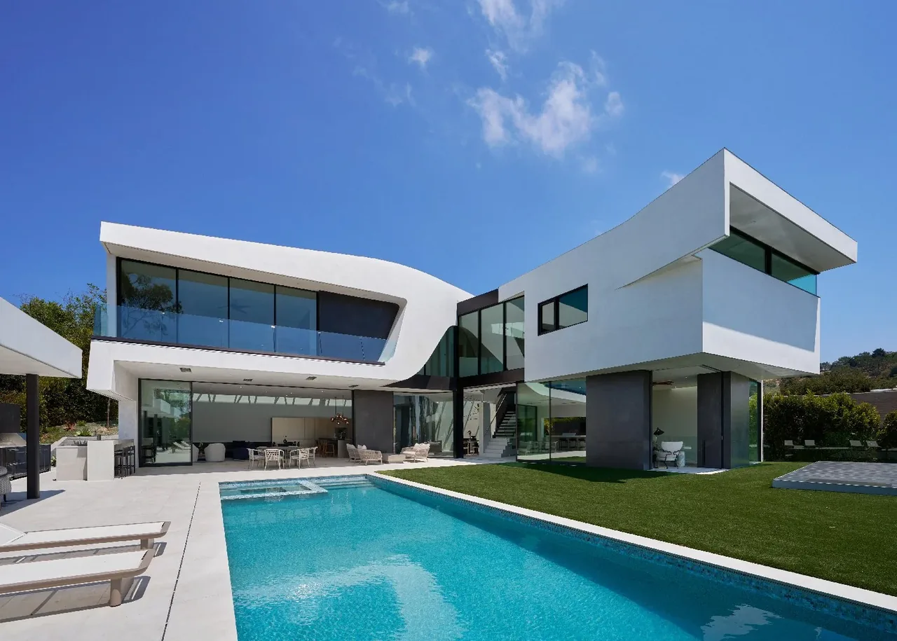 Modern white two-story house with cantilevered upper floor and large glass windows next to a blue swimming pool under a clear sky.