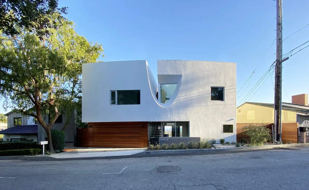 Modern white two-story house with cubic forms, a curved central depression, and wooden garage door on a sloped street.
