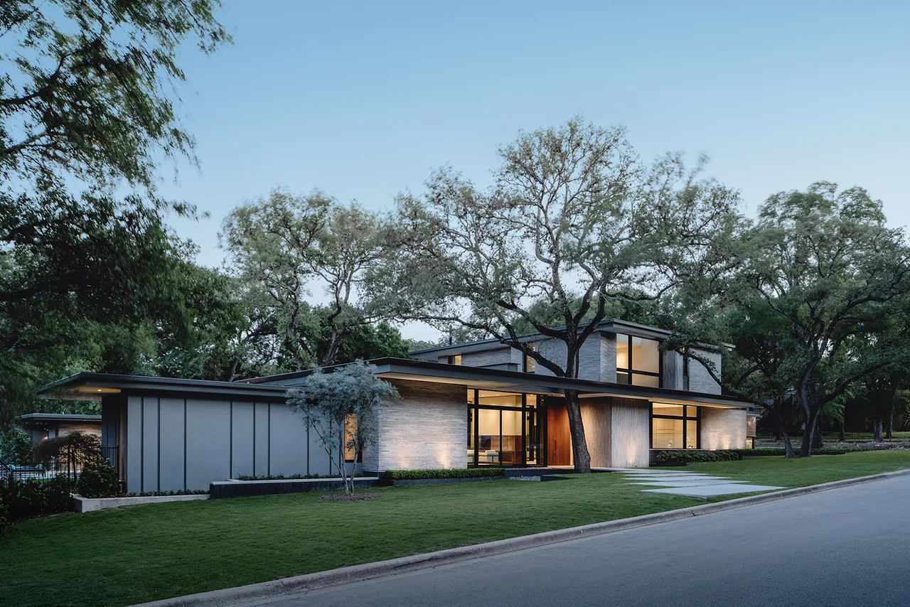 Modern two-story house exterior surrounded by trees at twilight with lit windows and gray siding.