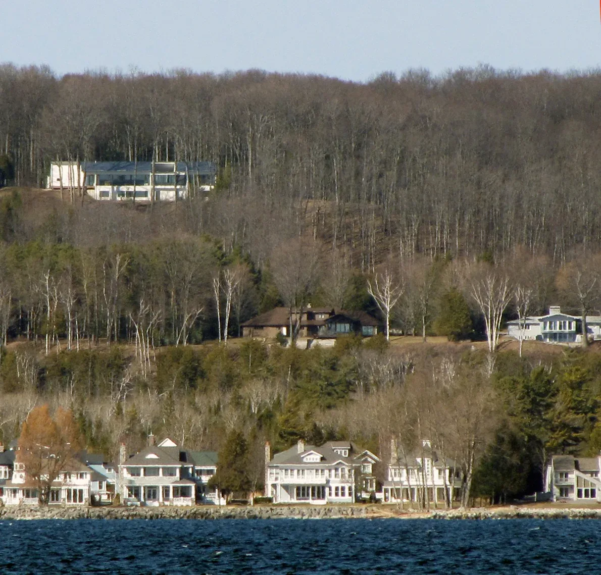 Modern house on a forested bluff overlooking a lake with traditional waterfront homes below.