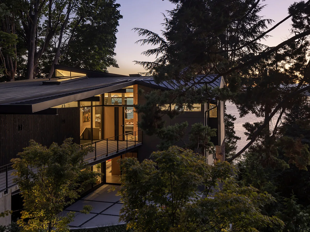 Exterior view of a modern two-story house at dusk with large windows and a metal shed roof surrounded by trees.