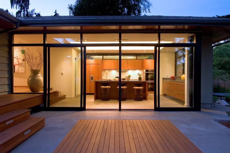 Evening view of a modern kitchen through large floor-to-ceiling glass doors with black frames and wooden decking outside.