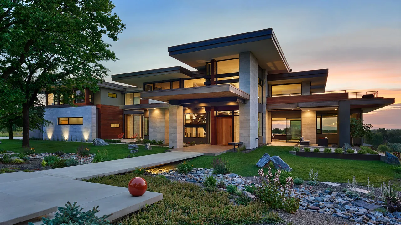Contemporary multi-level house exterior at dusk with large windows and flat roofs surrounded by landscaping.