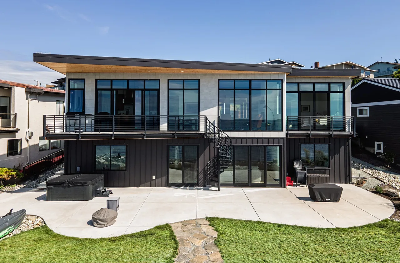 Rear exterior of a modern two-story house with large black-framed windows, upper balcony, and a spiral staircase to the lower patio.