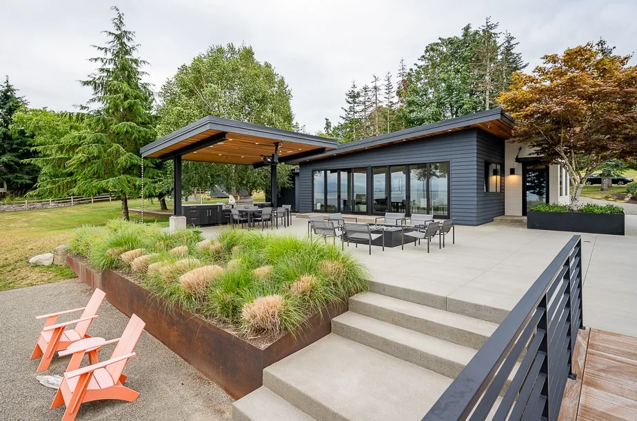 Modern house with dark exterior siding, large glass doors, and a covered patio with outdoor seating surrounded by greenery.