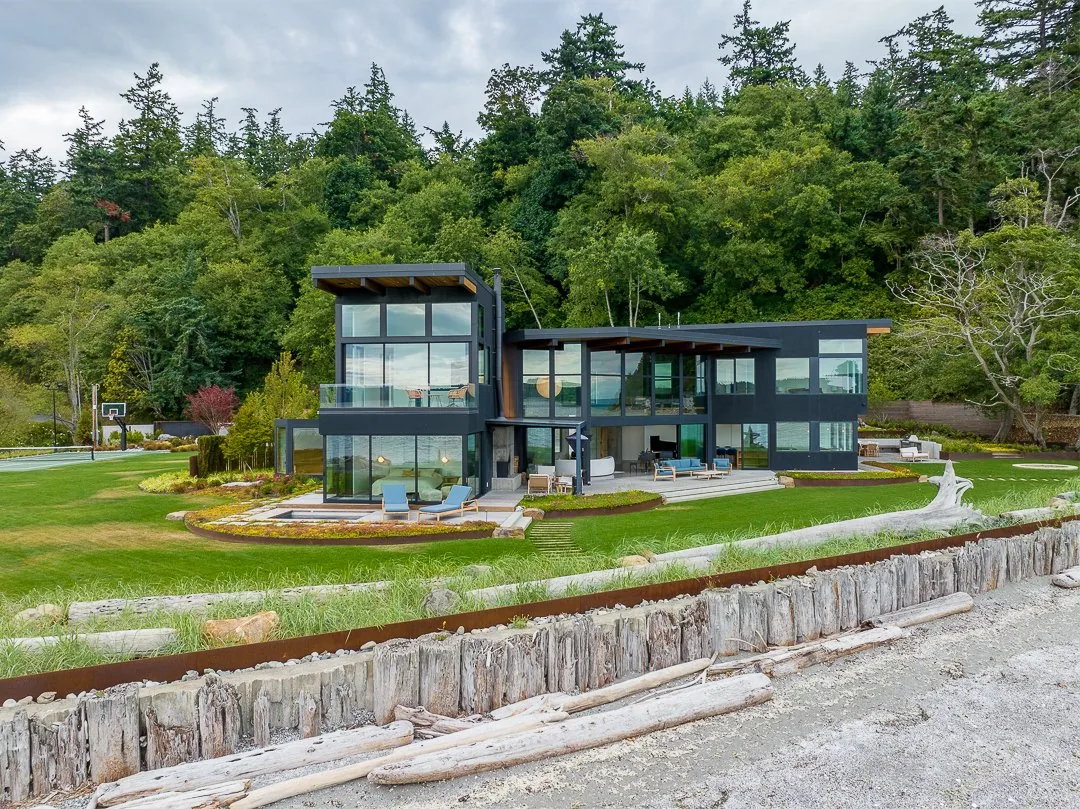 Modern two-story dark house with large floor-to-ceiling windows near a grassy lawn and rocky shoreline with driftwood.