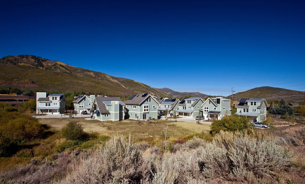 Cluster of light blue cottages with gray roofs on a flat plain against a mountainous backdrop under a clear blue sky.