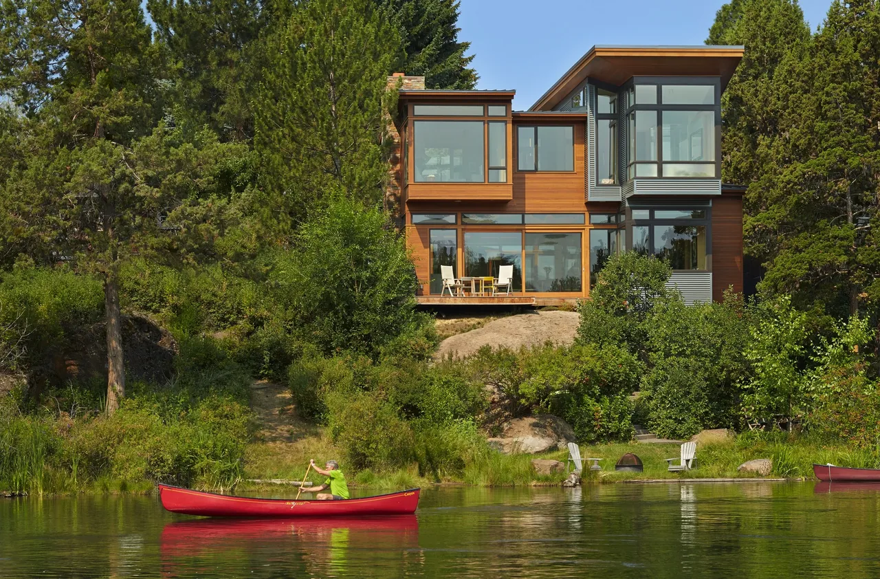 Modern two-story house with large windows and wood siding overlooking a lake with a person paddling a red canoe.