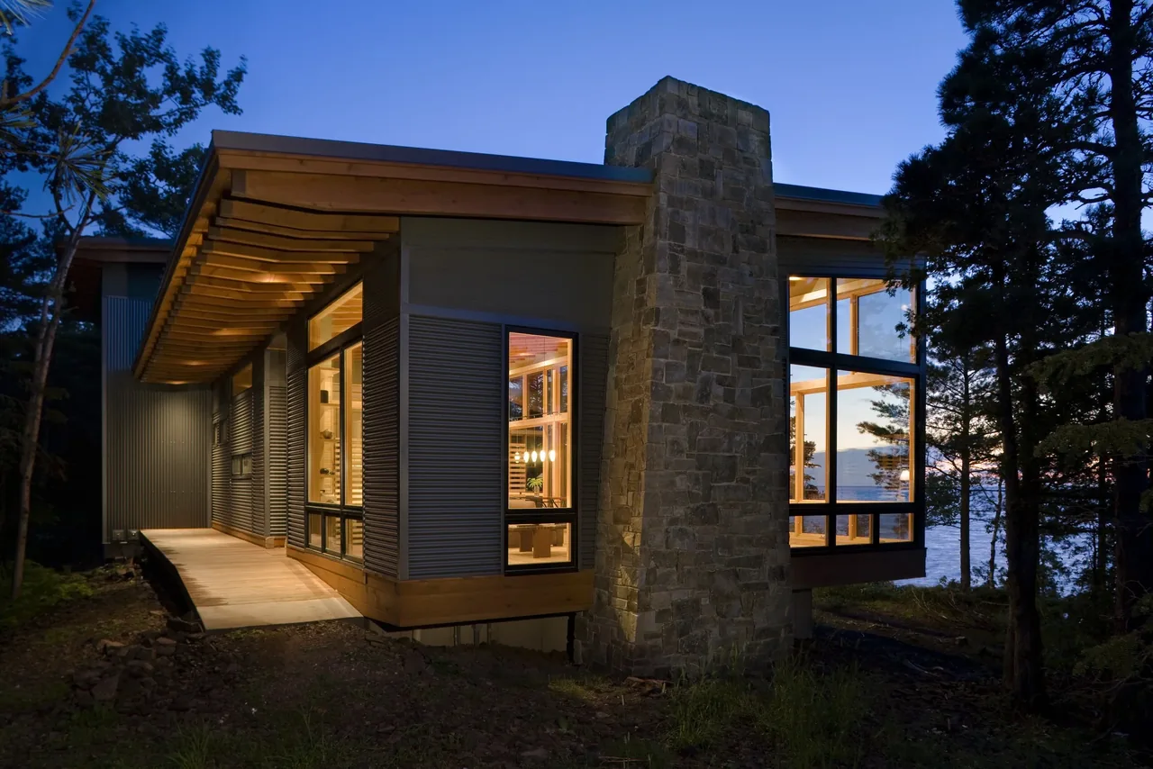 Exterior view of a modern house at dusk with large windows and a stone chimney near a wooded area and water.