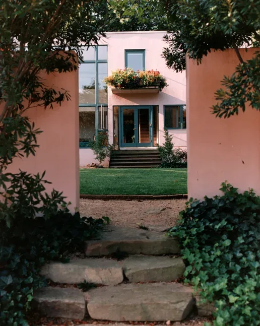 View through a garden opening with stone steps leading to a pink stucco house with green-framed doors and windows and a flower box.