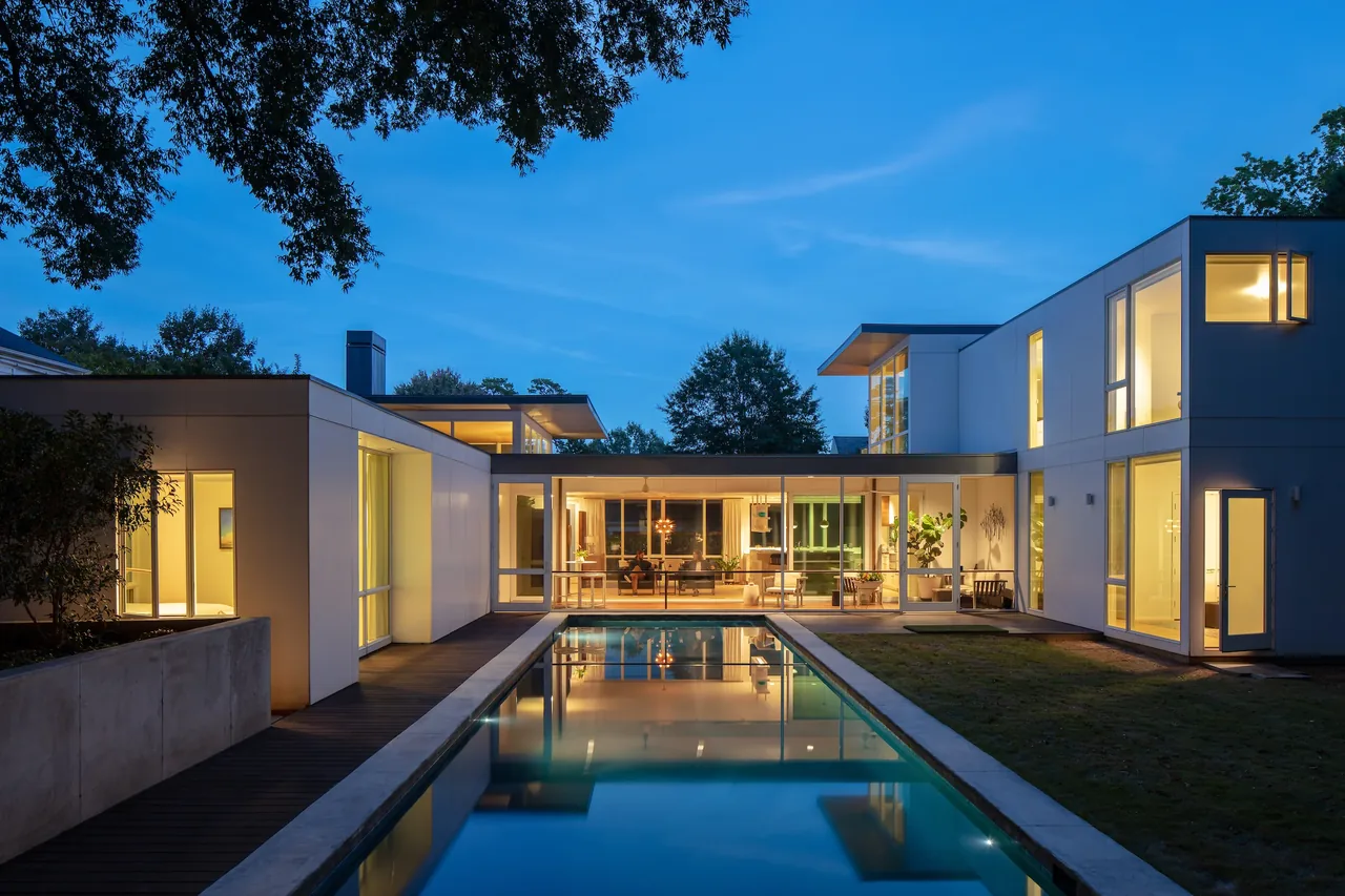 Modern house exterior at dusk with large glass windows and a rectangular swimming pool.