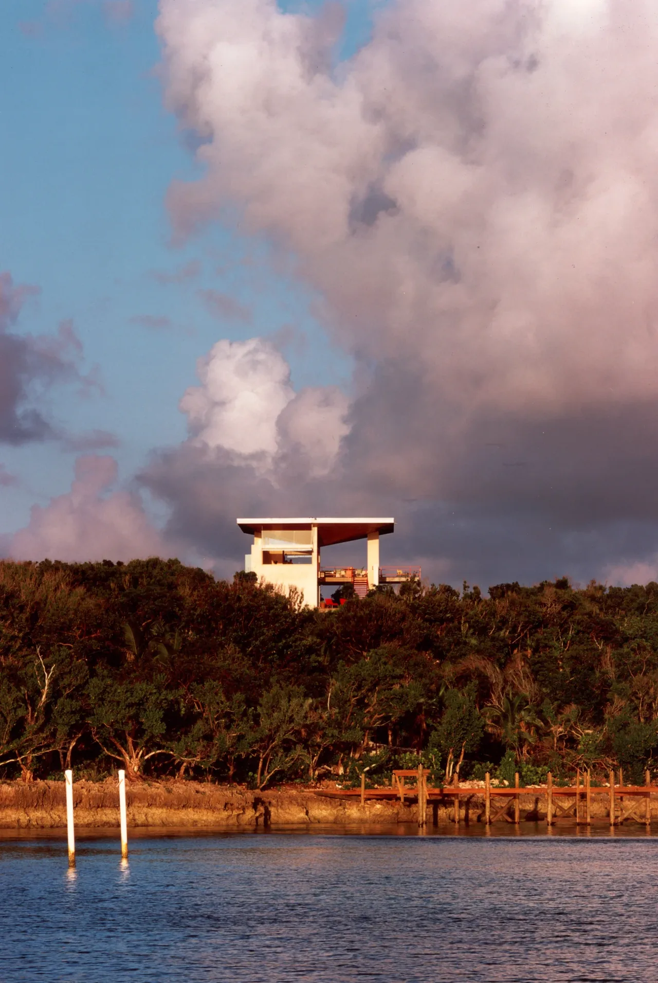 Modern vacation home with flat roof and open upper deck above greenery near a body of water under a cloudy sky.
